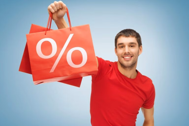Hombre sonriente con bolsas de compra rojas con símbolo de porcentaje, representando promociones y descuentos de temporada en Elektra.